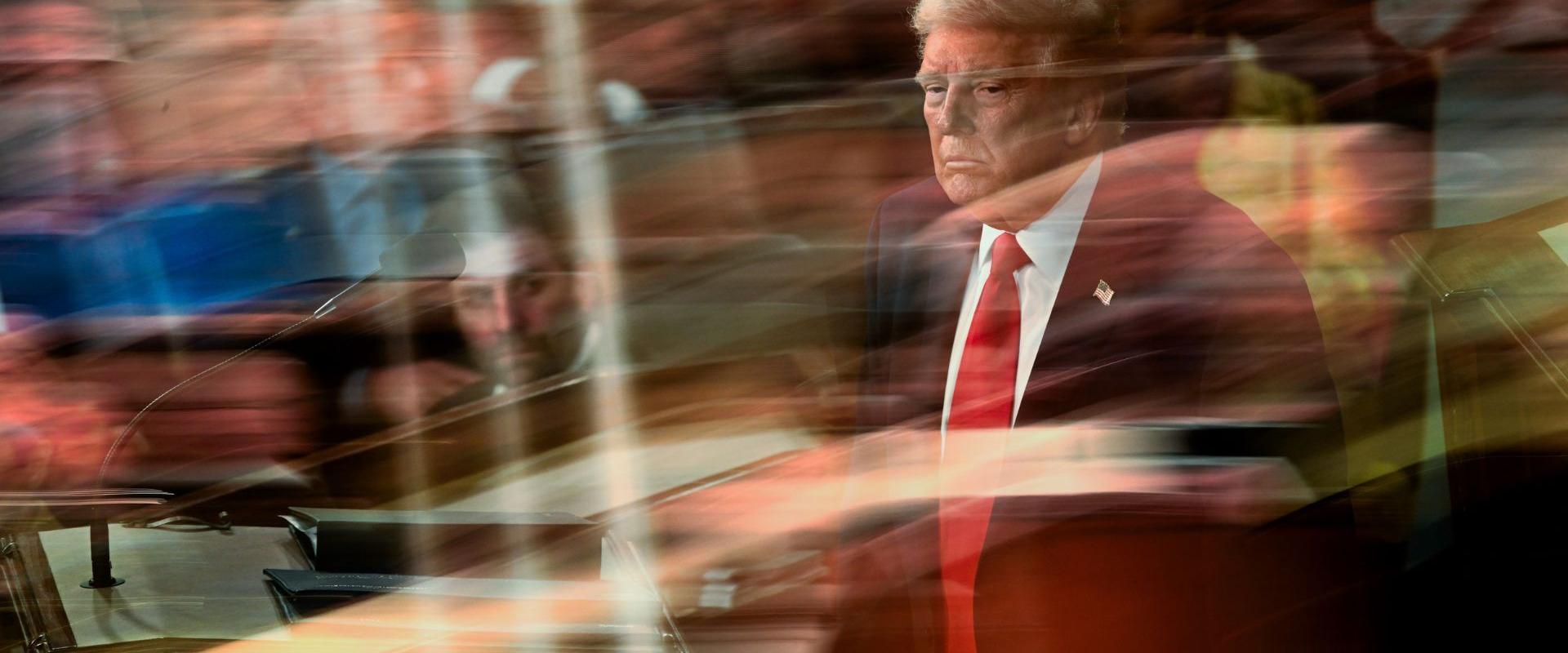 President Trump stands at the podium during the State of the Union address, viewed through a reflective glass surface that layers crowd reflections over his image. He wears a dark suit, red tie, and American flag lapel pin.